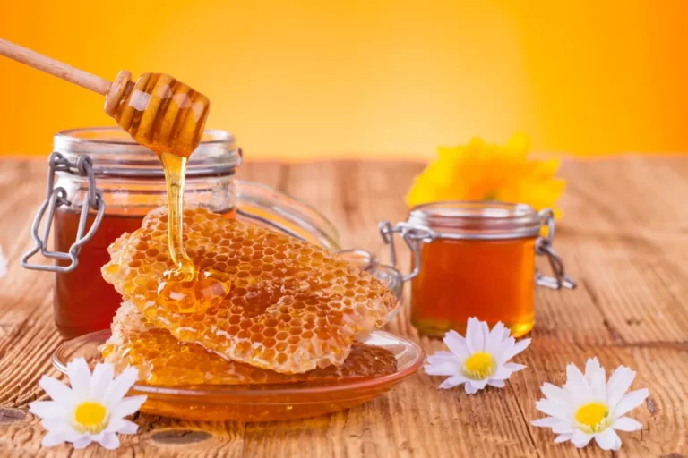 דבש ניגר בחלת דבש המונחת על צלחת ולצידה שתי צנצנות דבש על שולחן עץ